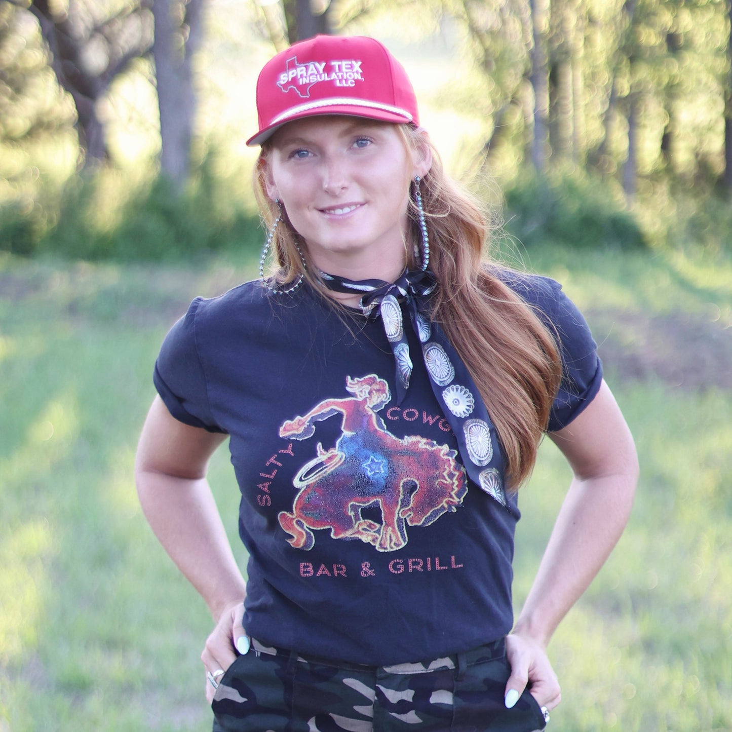 Woman wearing a navy t-shirt with a graphic design, camouflage pants, and a red cap in an outdoor setting.
