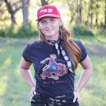 Load image into Gallery viewer, Woman wearing a navy t-shirt with a graphic design, camouflage pants, and a red cap in an outdoor setting.
