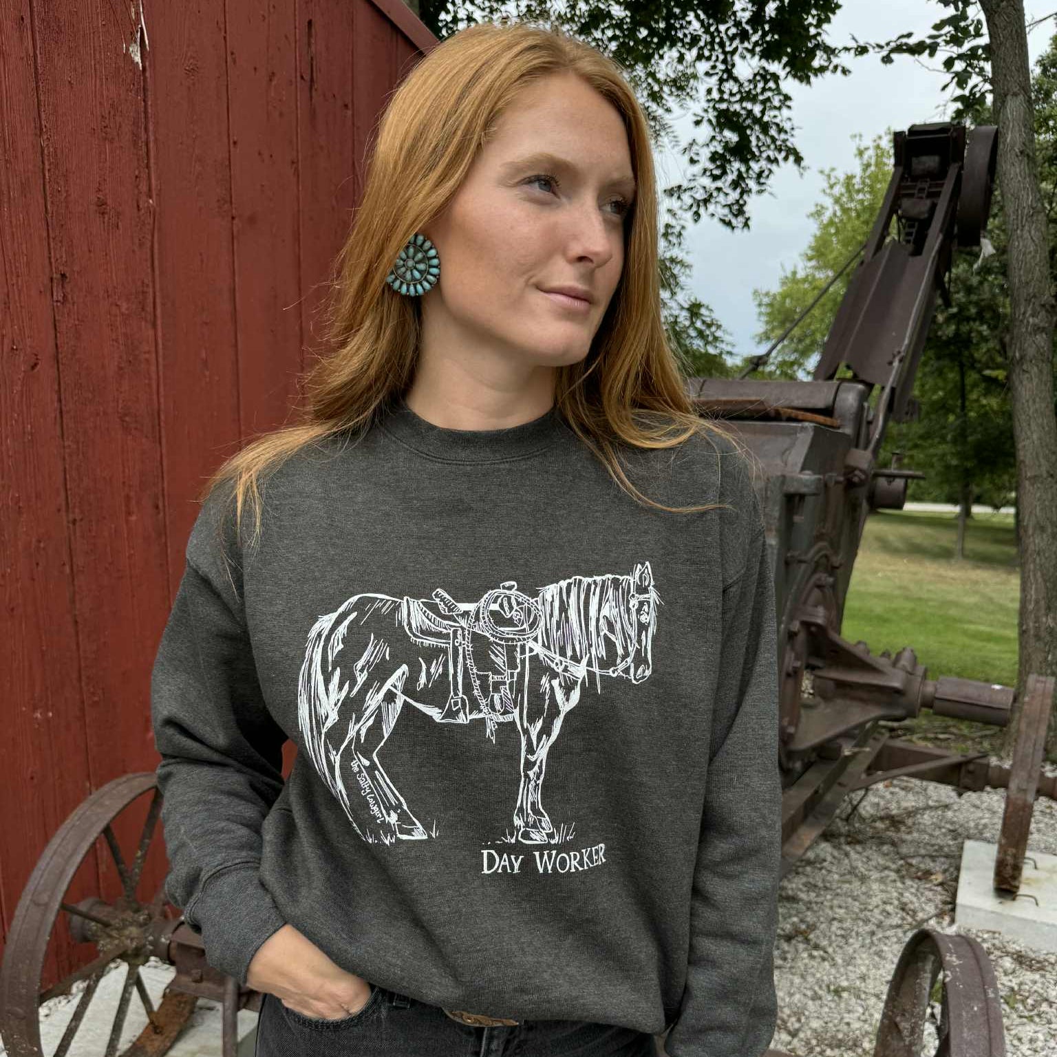 Woman wearing a dark gray sweatshirt with a horse graphic and text, standing in front of a red barn and old wagon.