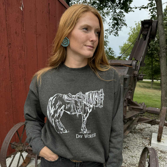 Woman wearing a dark gray sweatshirt with a horse graphic and text, standing in front of a red barn and old wagon.