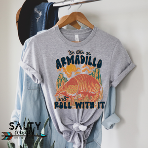 Hanging grey tee with southwest color design featuring an armadillo with the phrase "Be like an armadillo and roll with it.