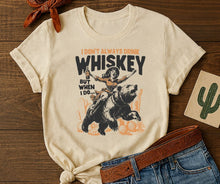 Load image into Gallery viewer, Beige t-shirt with graphic design of a person riding a bear and text, placed on a wooden surface with jeans and a belt.
