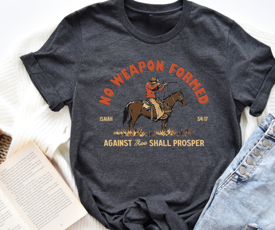 A dark heather gray graphic t-shirt featuring a western design with a cowboy on a horse and the text 'NO WEAPON FORMED AGAINST HEAR SHALL PROSPER', styled in a flat lay with a book and denim jeans on a white textured blanket.