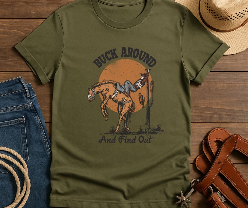 Green t-shirt with a graphic of a person on a horse and text 'Buck Around and Find Out' on a wooden surface with jeans, a hat, and leather strap.