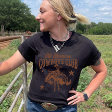 Load image into Gallery viewer, Blonde cowgirl standing next to arena fence. Wearing black tee with Cowboys Club logo
