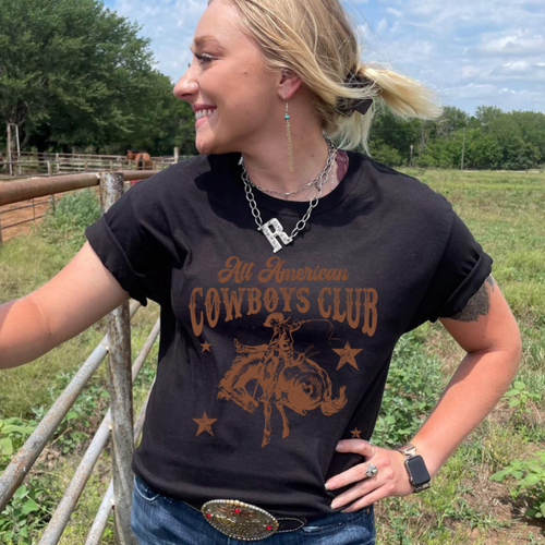 Blonde cowgirl standing next to arena fence. Wearing black tee with Cowboys Club logo