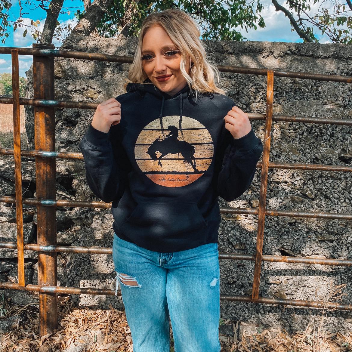 Blonde cowgirl wearing a black hoodie featuring a bucking horse design. 